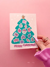 Load image into Gallery viewer, A hand holding a pink Christmas greeting card featuring a teal decorated Christmas tree with ornaments and the words 'MERRY CHRISTMAS' in pink, set against a solid pink background.
