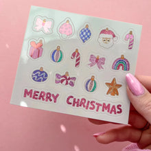 Load image into Gallery viewer, A hand with pink manicured nails holds a sheet of Christmas-themed stickers against a solid pink background. The stickers include illustrations of bows, ornaments, candy canes, a Santa face, a rainbow, a gift box, and a star, with 'MERRY CHRISTMAS' written at the bottom in pink glitter-style lettering.