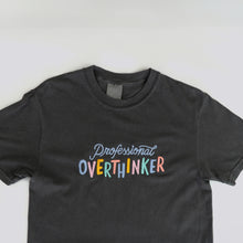Load image into Gallery viewer, A graphite grey, 100% cotton t-shirt with a playful, embroidered message that reads &quot;Professional Overthinker&quot; in colorful, handwritten-style lettering. The shirt has a relaxed, unisex fit, perfect for everyday wear.