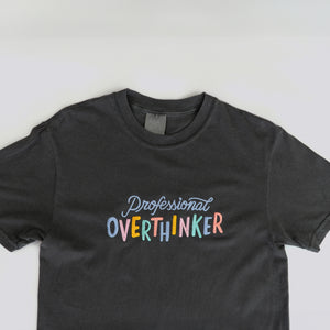 A graphite grey, 100% cotton t-shirt with a playful, embroidered message that reads "Professional Overthinker" in colorful, handwritten-style lettering. The shirt has a relaxed, unisex fit, perfect for everyday wear.