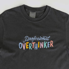 Load image into Gallery viewer, A graphite grey, 100% cotton t-shirt with a playful, embroidered message that reads &quot;Professional Overthinker&quot; in colorful, handwritten-style lettering. The shirt has a relaxed, unisex fit, perfect for everyday wear.