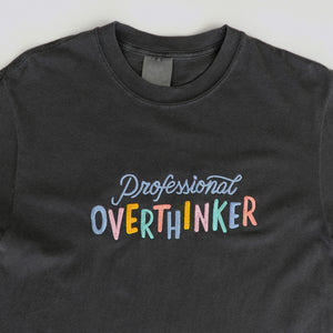 A graphite grey, 100% cotton t-shirt with a playful, embroidered message that reads "Professional Overthinker" in colorful, handwritten-style lettering. The shirt has a relaxed, unisex fit, perfect for everyday wear.