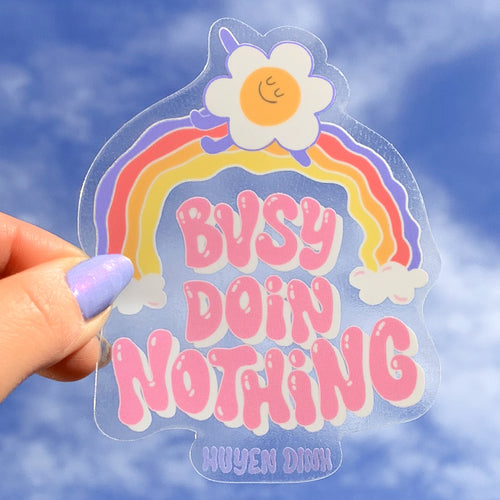 A clear vinyl sticker featuring a playful rainbow with a smiling sun flower on top. The phrase 