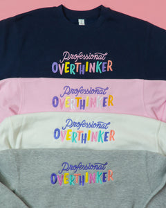 Four sweatshirts stacked on top of each other. The top sweatshirt is navy, the second is pink, the third is white, and the bottom is gray. All four sweatshirts feature the embroidered phrase "Professional Overthinker" in different color combinations. The sweatshirts are made of a comfortable blend of 80% cotton and 20% polyester and are designed to be both relatable and humorous, perfect for anyone who enjoys a good laugh while acknowledging their tendency to overthink.