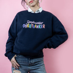 A navy sweatshirt with the embroidered phrase "Professional Overthinker" in a playful rainbow of colors. The sweatshirt is made of a comfortable blend of 80% cotton and 20% polyester and has a unisex fit. The design is both relatable and humorous, making it a great choice for anyone who enjoys a good laugh while acknowledging their tendency to overthink.